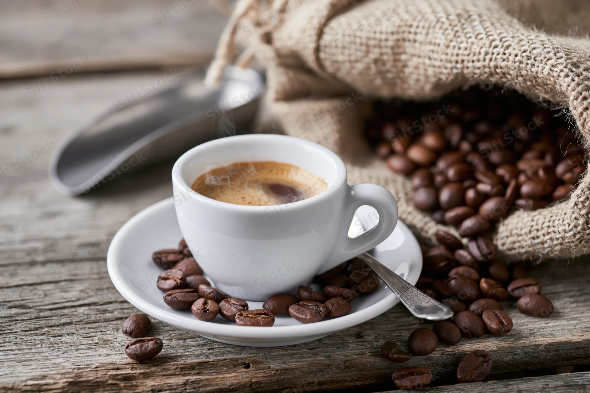 White ceramic espresso cup with crema and steam on saucer surrounded by roasted coffee beans on wooden table