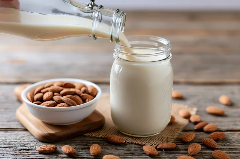 Almond milk being poured from a glass bottle into a jar on a wooden table with scattered almonds and a bowl nearby