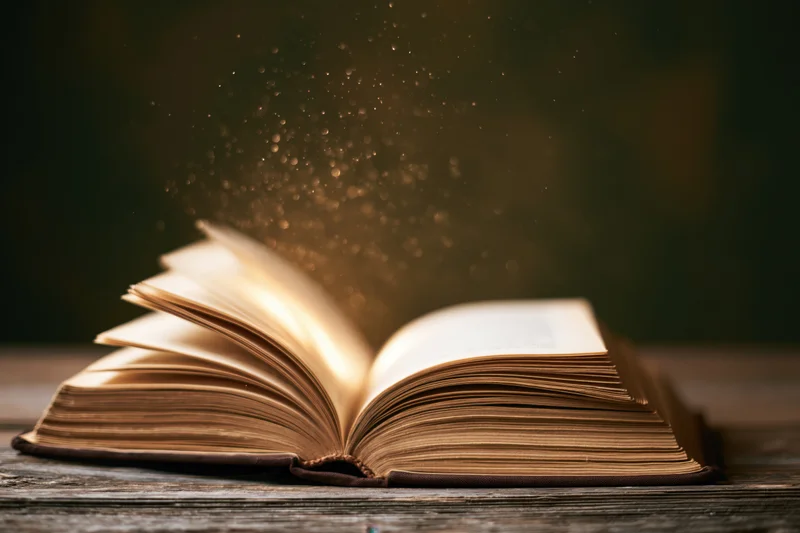 Open old hardcover book on worn wooden table with glowing dust particles rising from pages