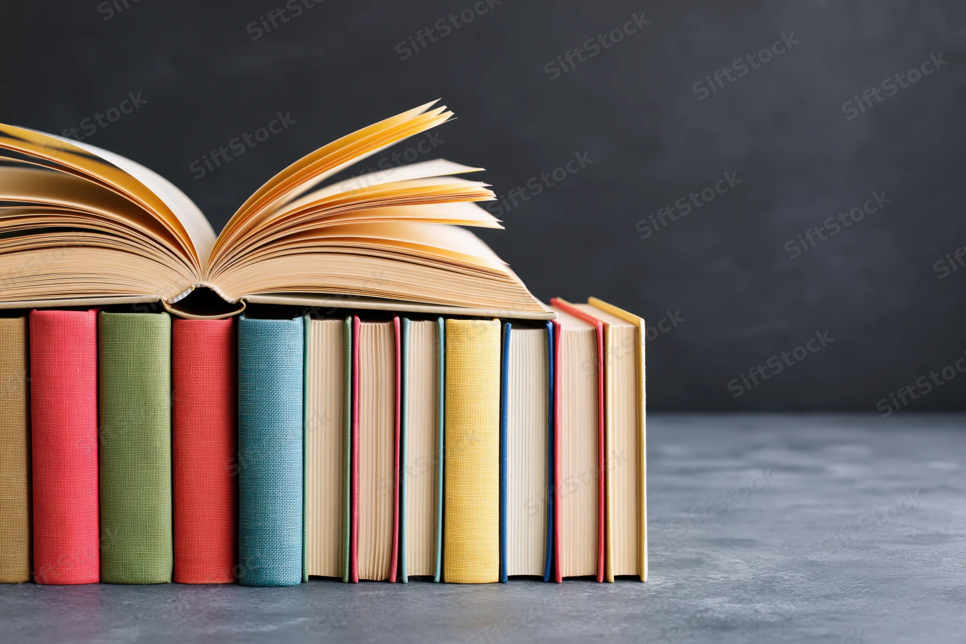 Open hardcover book with fanned yellowed pages resting on a stack of colorful books against a gray background