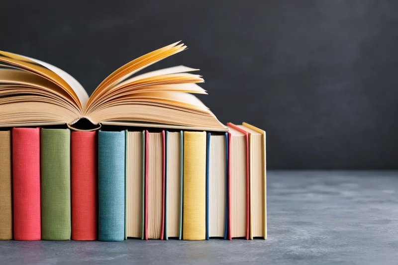 Open hardcover book with fanned yellowed pages resting on a stack of colorful books against a gray background