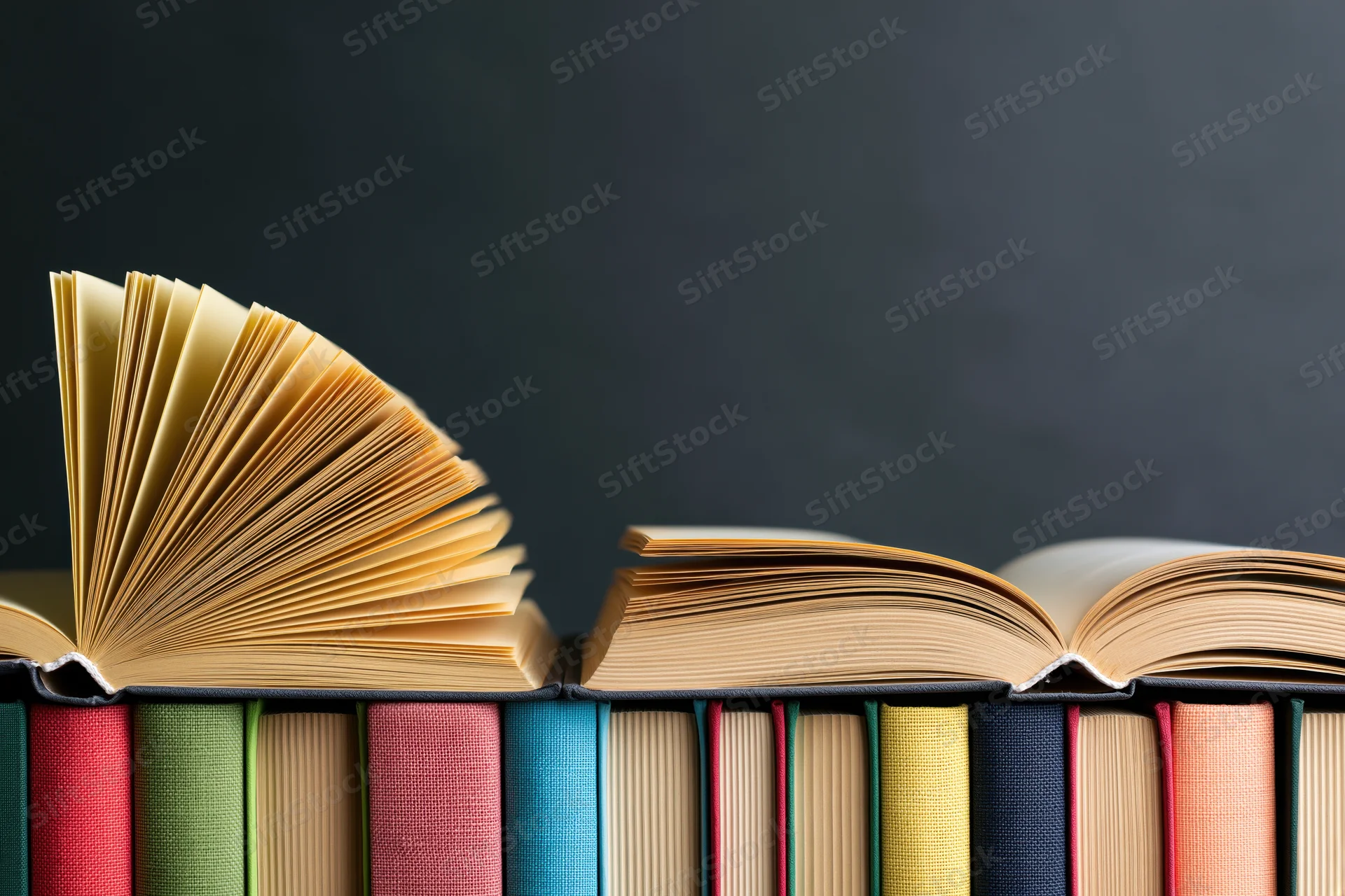 Open hardcover books with fanned yellowed pages resting on a row of colorful spines against a dark background