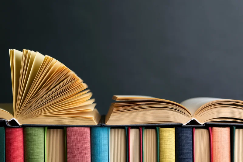 Open hardcover books with fanned yellowed pages resting on a row of colorful spines against a dark background