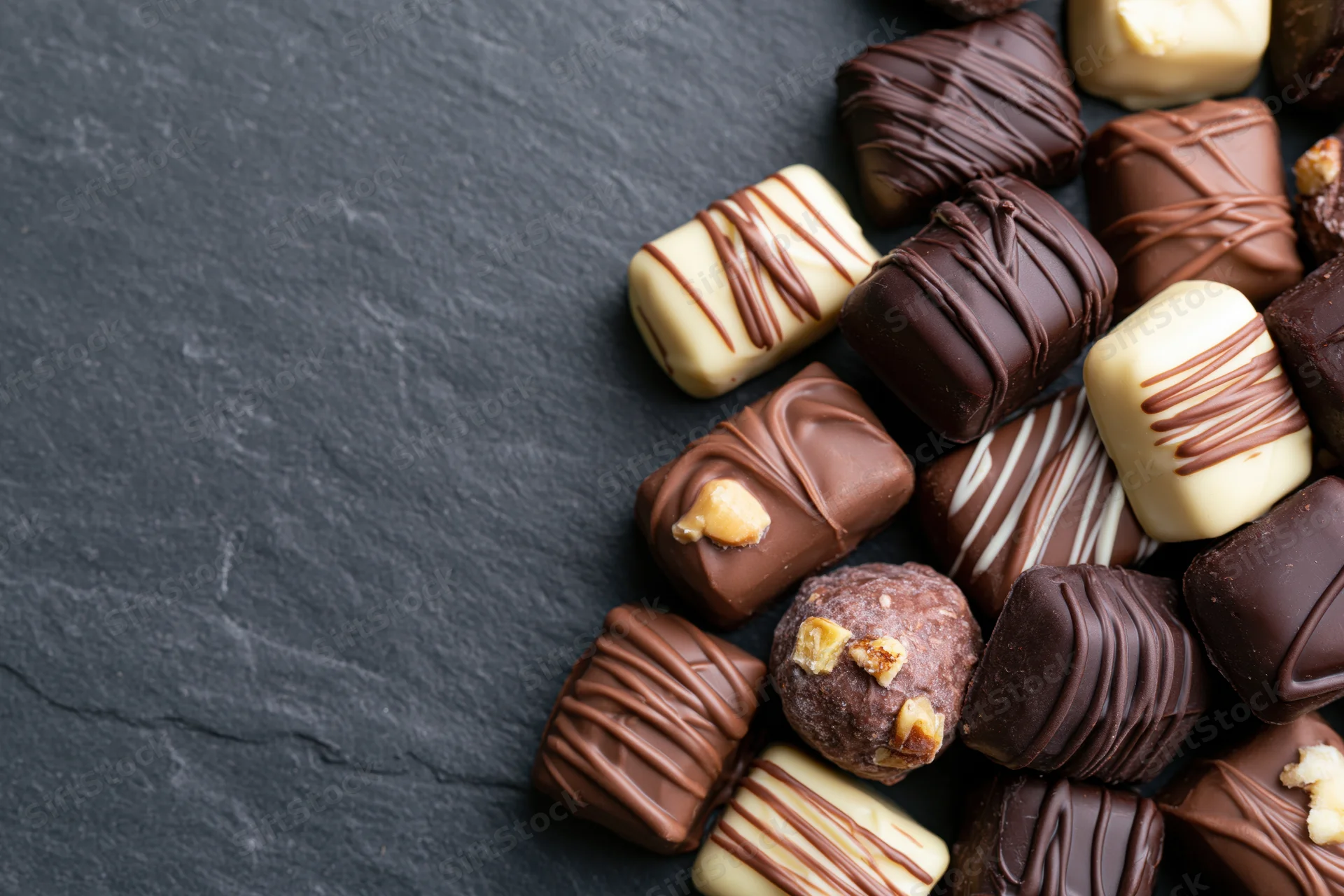 Assorted luxury chocolate pralines and truffles scattered on a dark slate surface with copyspace on the left