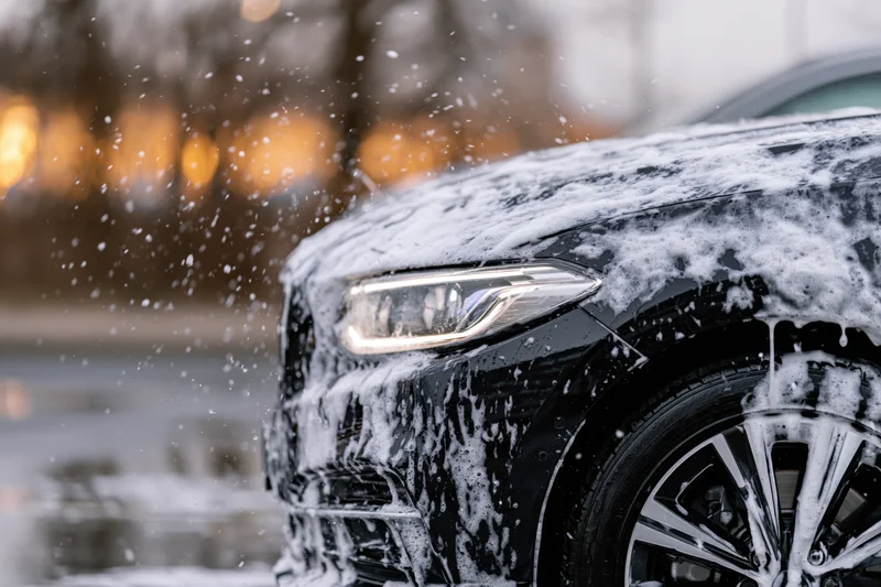 Black luxury sedan covered in white soap foam during outdoor car wash at night
