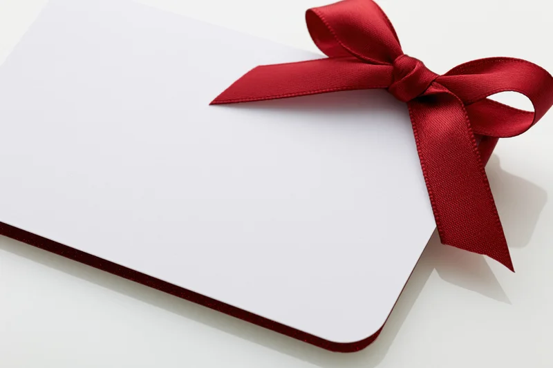 Blank white gift card with red satin ribbon bow on white surface