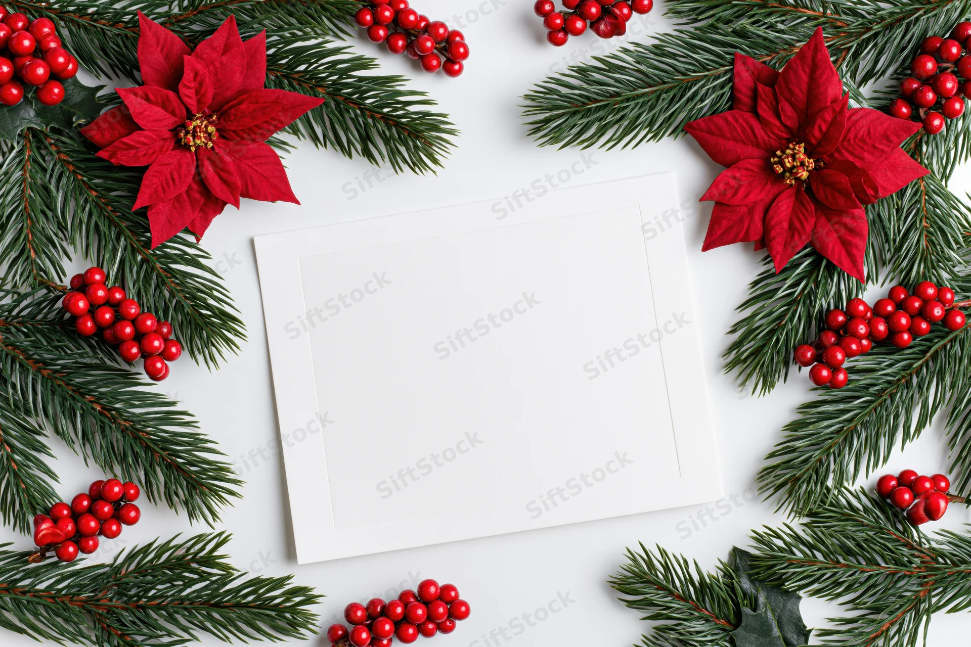 Blank white greeting card centered on a flat lay arrangement of poinsettias, evergreen branches and red berries
