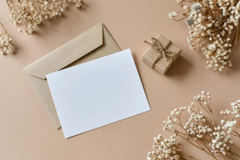 Blank white card with kraft envelope and dried flowers arranged on beige surface