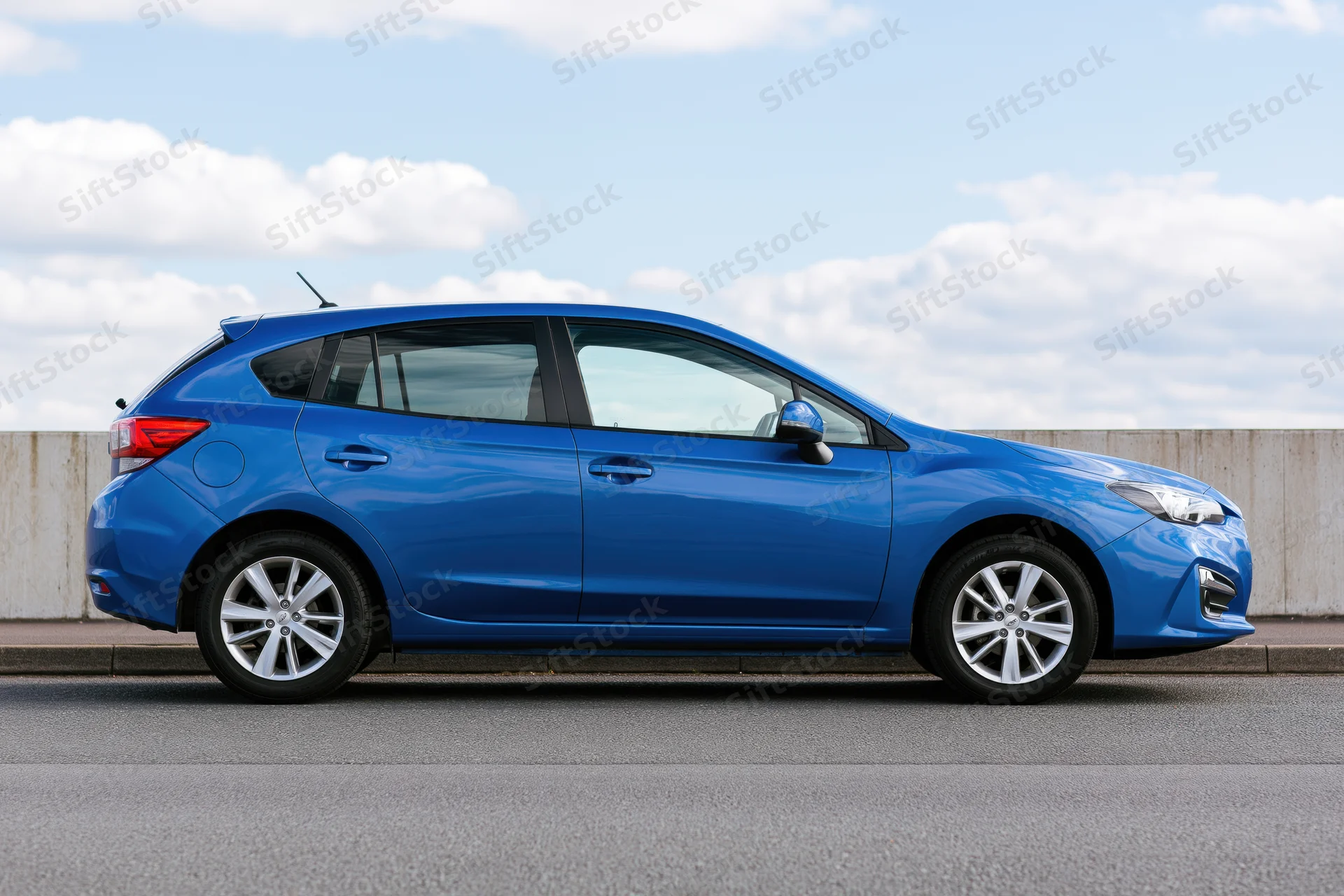 Blue compact hatchback parked on asphalt beside a low concrete barrier under a partly cloudy sky