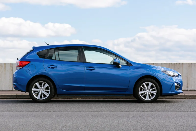 Blue compact hatchback parked on asphalt beside a low concrete barrier under a partly cloudy sky