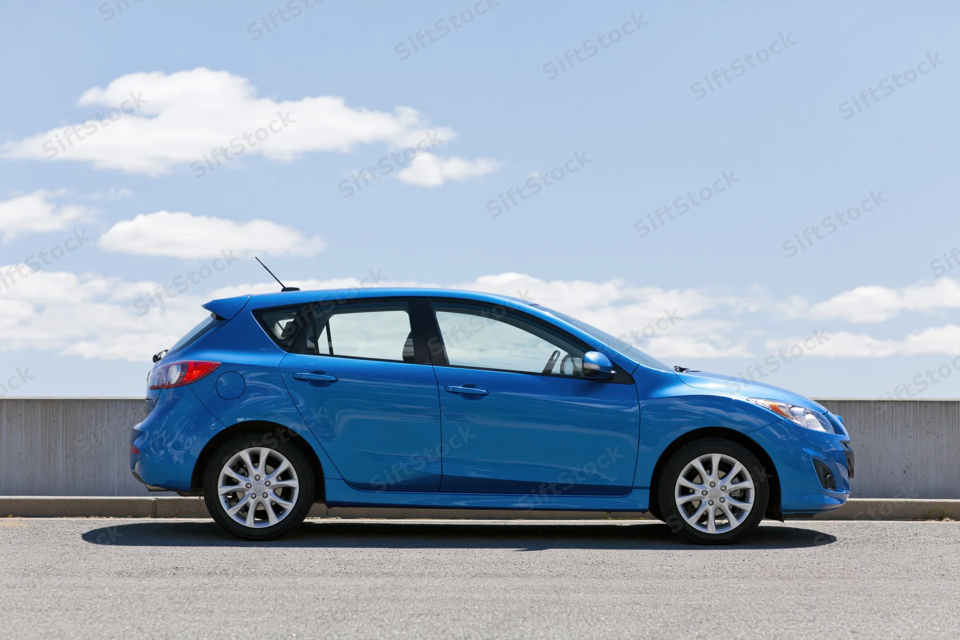 Blue compact hatchback parked on asphalt beside a low concrete wall under a partly cloudy sky