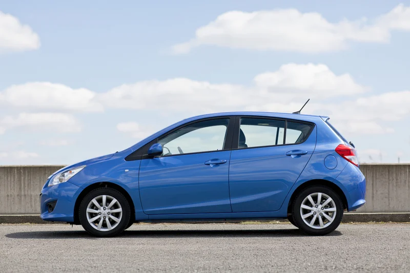 Blue compact hatchback parked on asphalt beside a low concrete wall under a partly cloudy sky