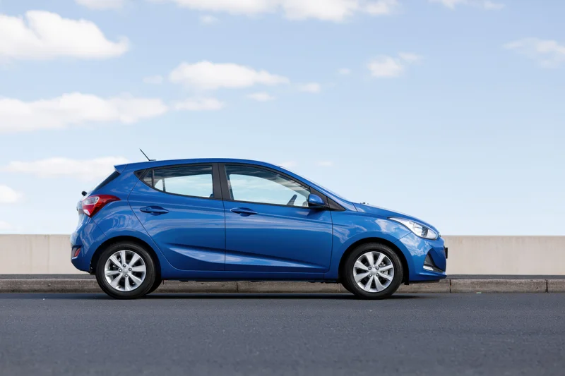 Blue compact hatchback parked on asphalt beside low concrete wall under clear sky