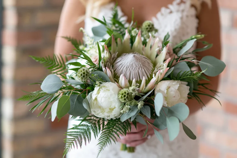 Bride holding a lush bridal bouquet with a large king protea in front of a brick wall