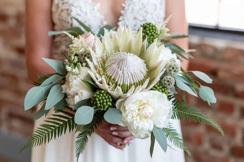 Bride holding a lush bridal bouquet with a large king protea in front of a brick wall