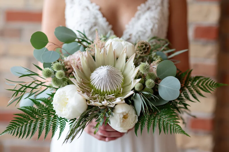 Bride holding a lush bridal bouquet with a large king protea in front of a brick wall
