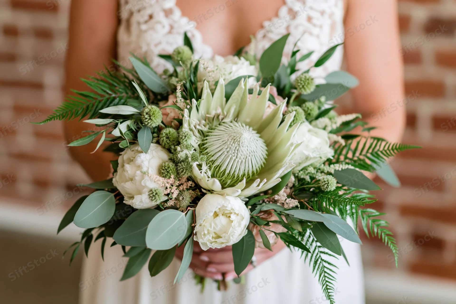 Bride holding a lush bridal bouquet with a large king protea in front of a brick wall