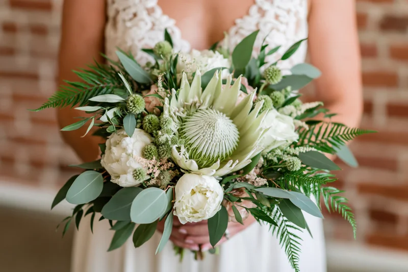 Bride holding a lush bridal bouquet with a large king protea in front of a brick wall