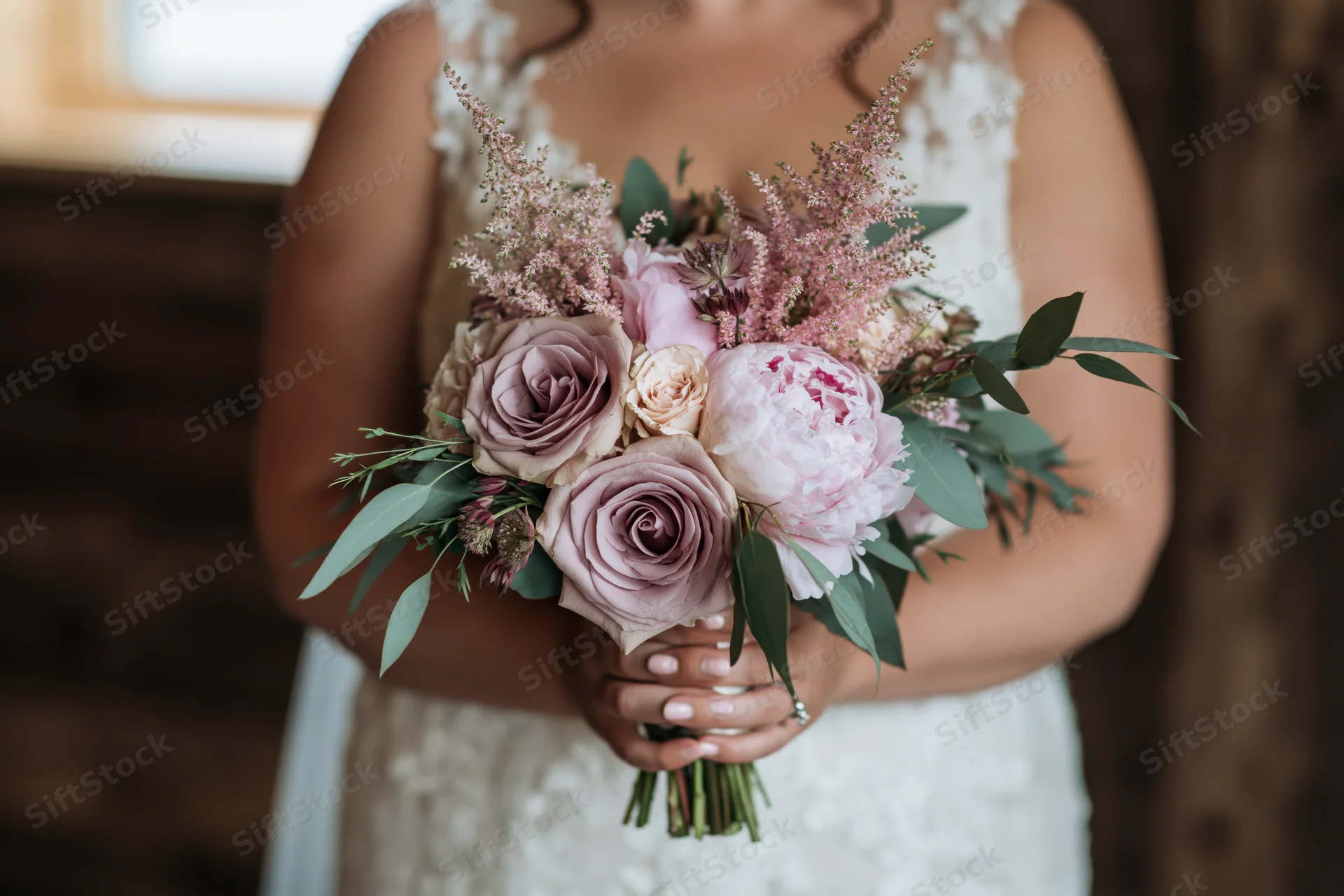 Bride holding a floral bouquet of dusty mauve roses and blush peonies with green foliage in a white lace dress