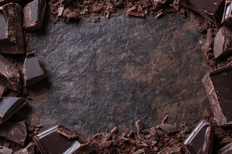 Broken dark chocolate chunks and shavings arranged in a frame around a dark textured surface with copyspace in center