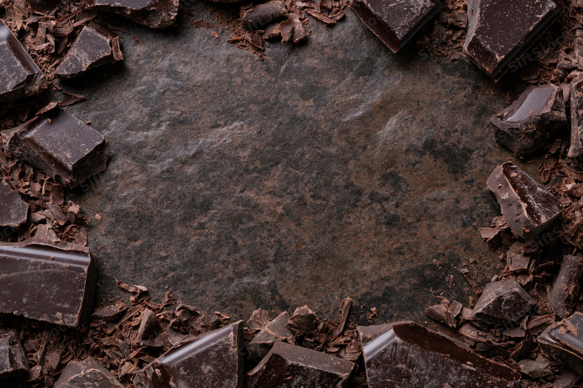 Broken dark chocolate chunks and shavings arranged in a frame around a slate surface with copyspace in center
