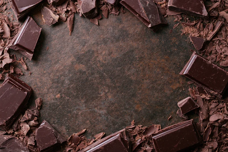 Broken dark chocolate chunks and shavings arranged in a frame around a dark textured surface with copyspace in center