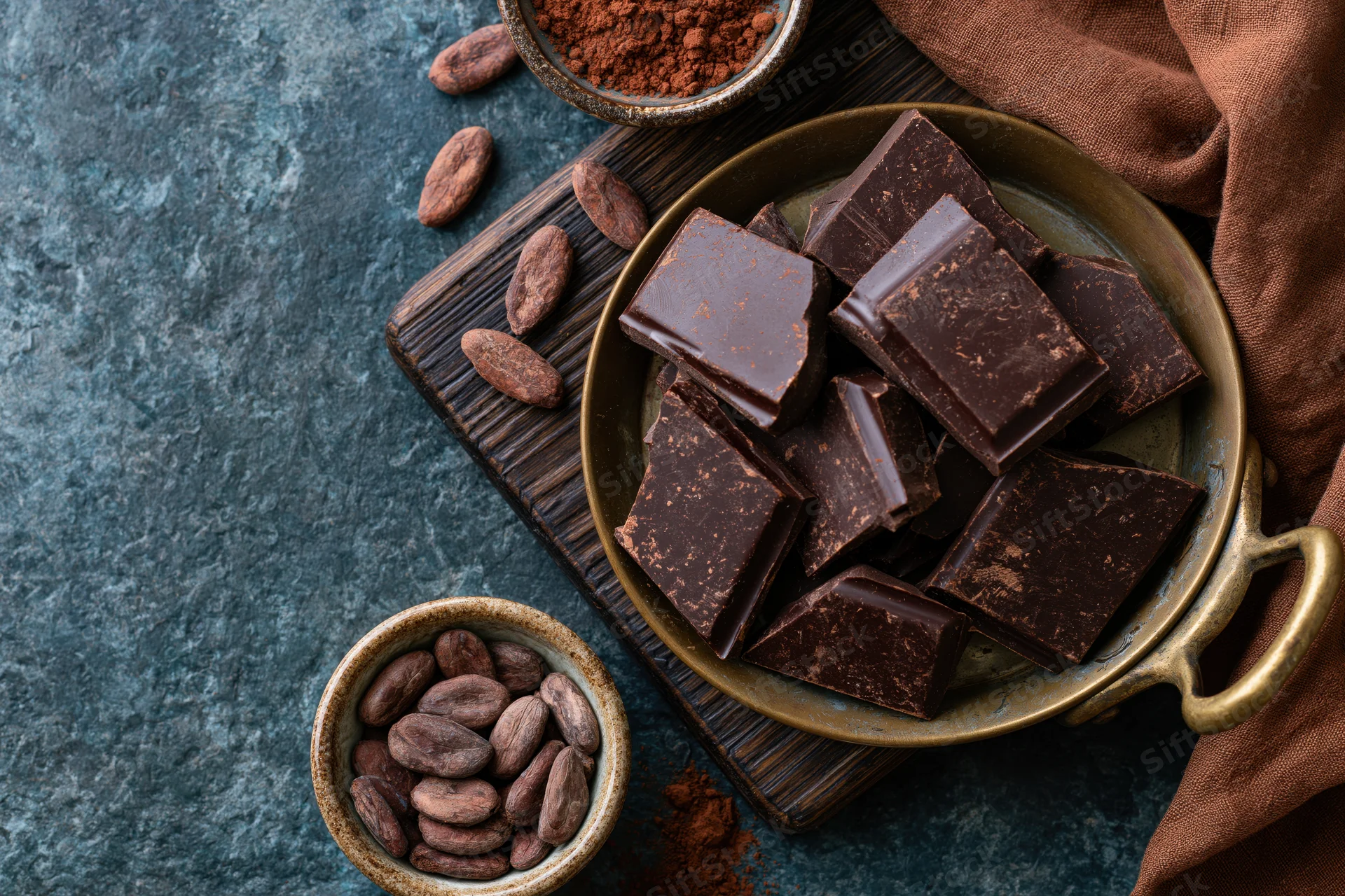 Broken dark chocolate chunks piled in a vintage brass pan on a wooden board with cocoa beans and powder nearby