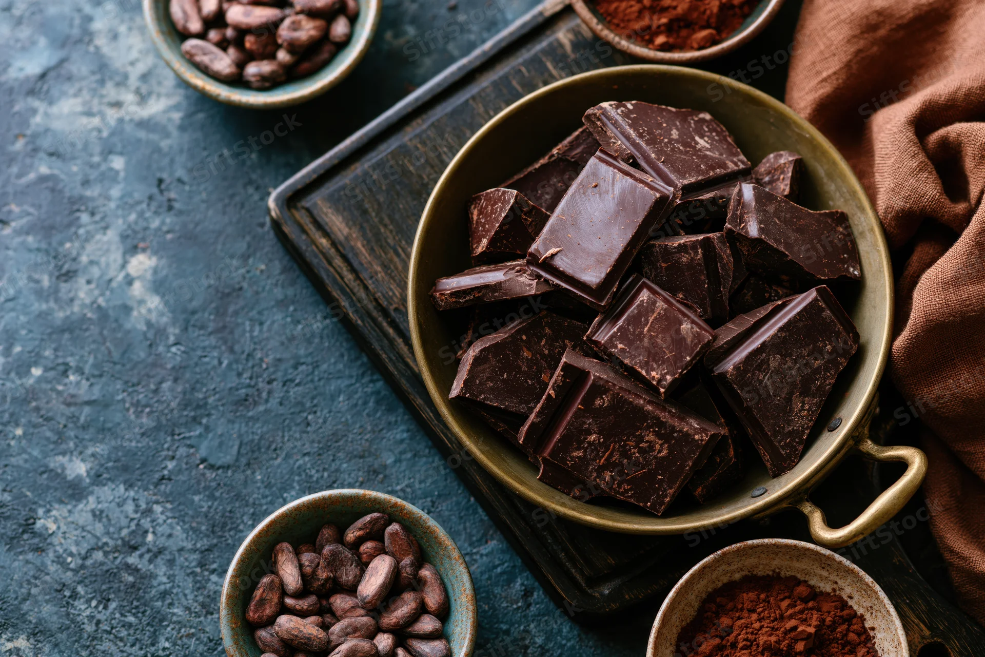 Broken dark chocolate chunks in a vintage brass pan on a rustic blue surface with cocoa beans and powder in small bowls nearby