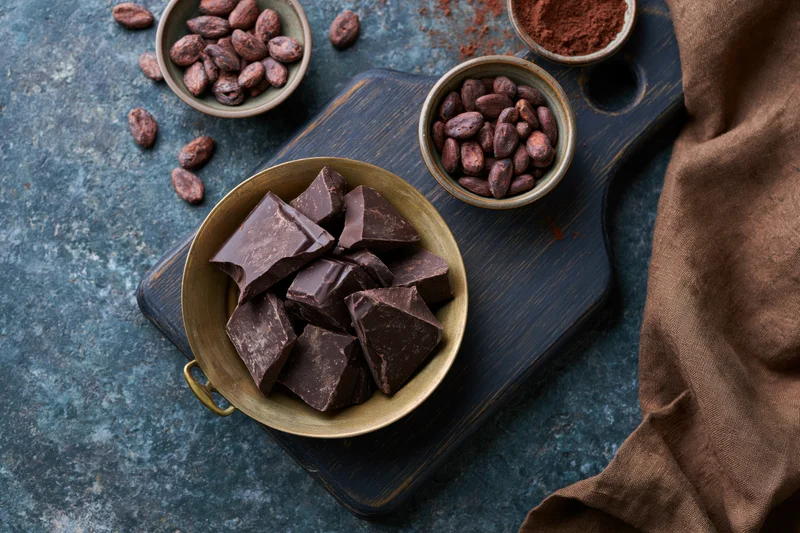 Broken dark chocolate chunks in a vintage brass pan on a wooden cutting board with cocoa beans and powder nearby