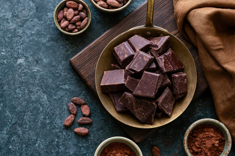 Broken dark chocolate chunks piled in a vintage brass pan on a wooden board with cocoa beans and powder around