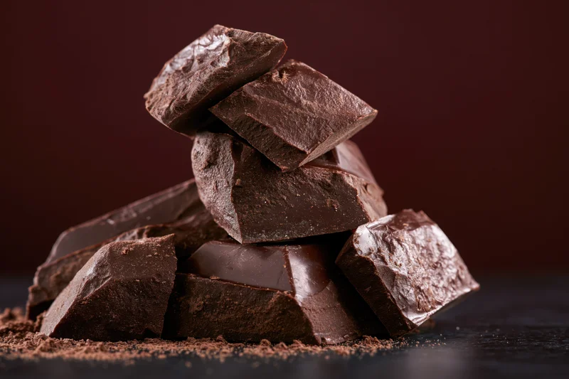 Broken dark chocolate chunks stacked into a rough tower closeup on dark surface with cocoa powder sprinkled around