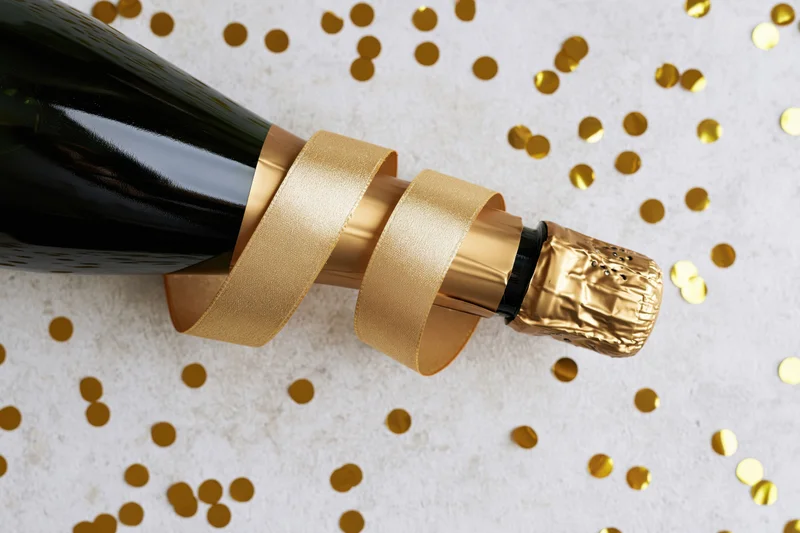 Champagne bottle lying on its side with curled gold ribbon and scattered confetti on light surface