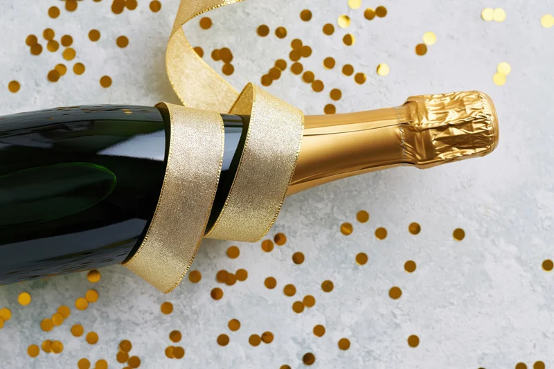 Champagne bottle lying on its side with a curled gold ribbon on a white textured surface scattered with gold confetti