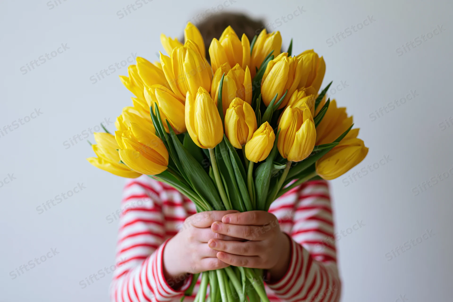 Child holding a large bouquet of yellow tulips in front of their face, wearing a red and white striped shirt