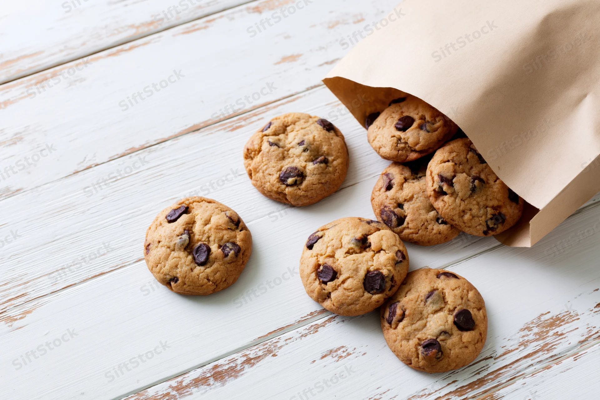 Chocolate chip cookies spilling from an open kraft paper bag onto a white wooden table