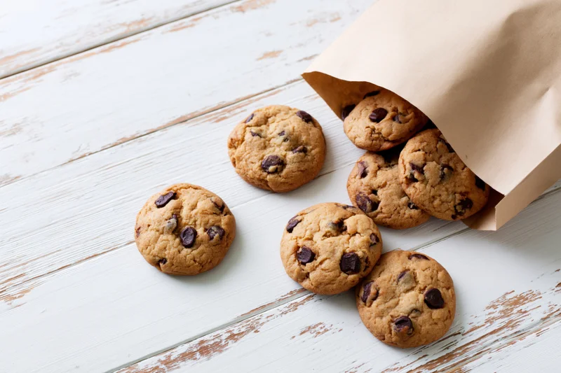 Chocolate chip cookies spilling from an open kraft paper bag onto a white wooden table