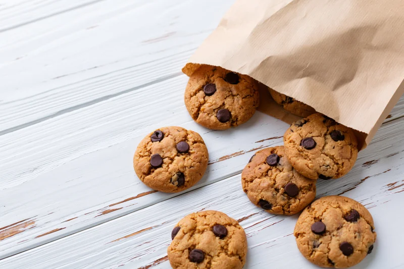 Chocolate chip cookies spilling from a kraft paper bag onto a white wooden tabletop