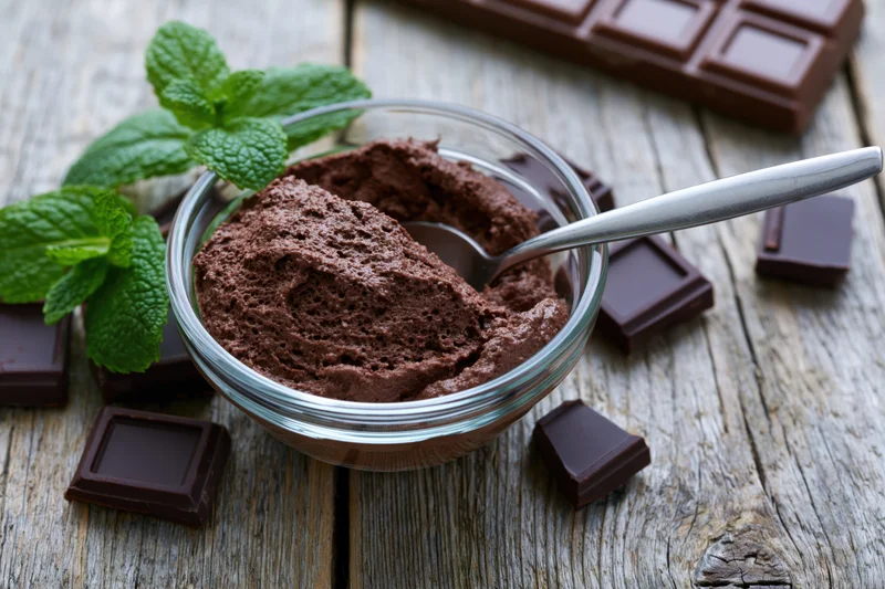 Chocolate mousse in a glass bowl with a metal spoon and mint leaves on a rustic wooden table