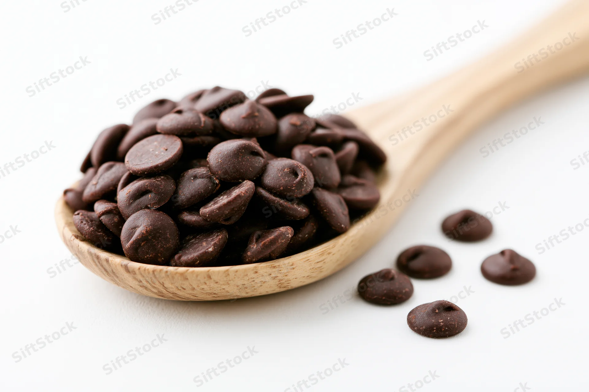 Dark chocolate chips piled in a wooden spoon with a few scattered beside it on a white surface