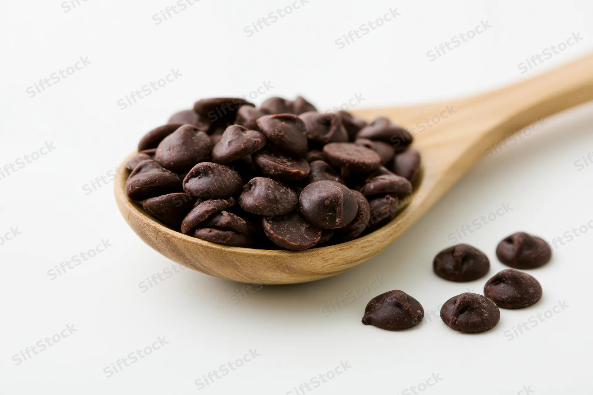Dark chocolate chips piled in a wooden spoon with a few scattered beside it on white background
