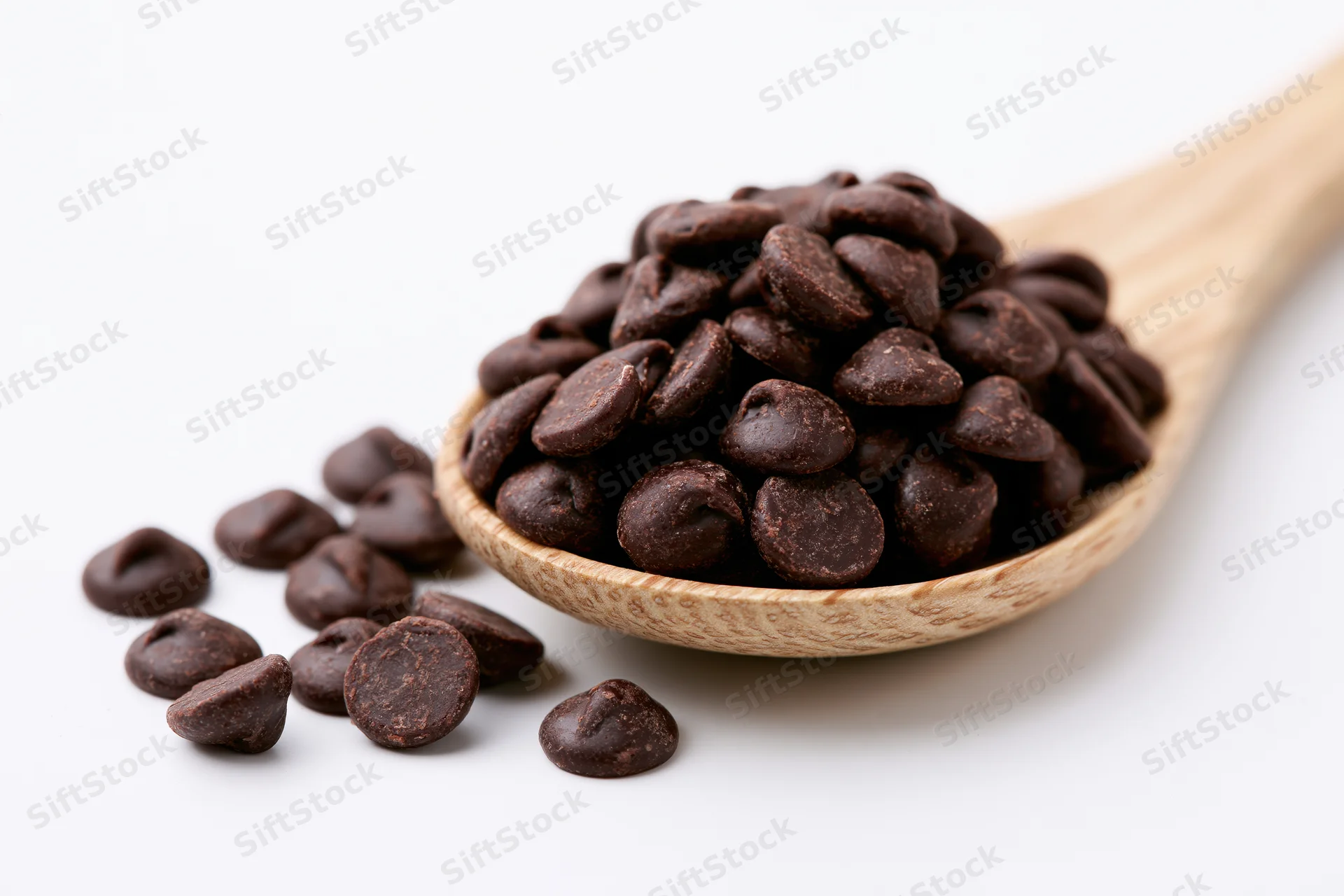 Dark chocolate chips piled in a wooden spoon with some scattered beside it on white background