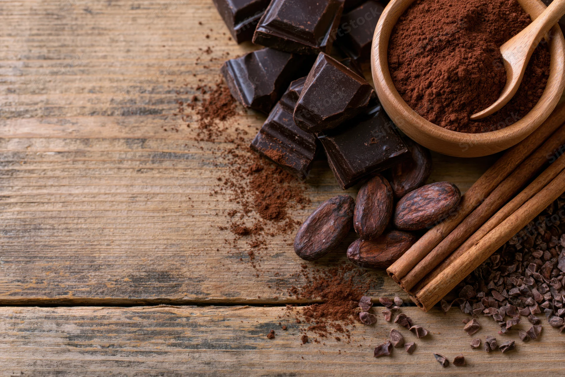 Dark chocolate chunks and cocoa powder in a wooden bowl with cinnamon sticks and cocoa beans on a rustic wood surface
