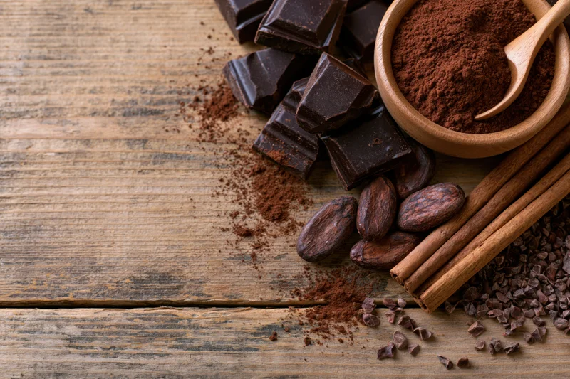 Dark chocolate chunks and cocoa powder in a wooden bowl with cinnamon sticks and cocoa beans on a rustic wood surface