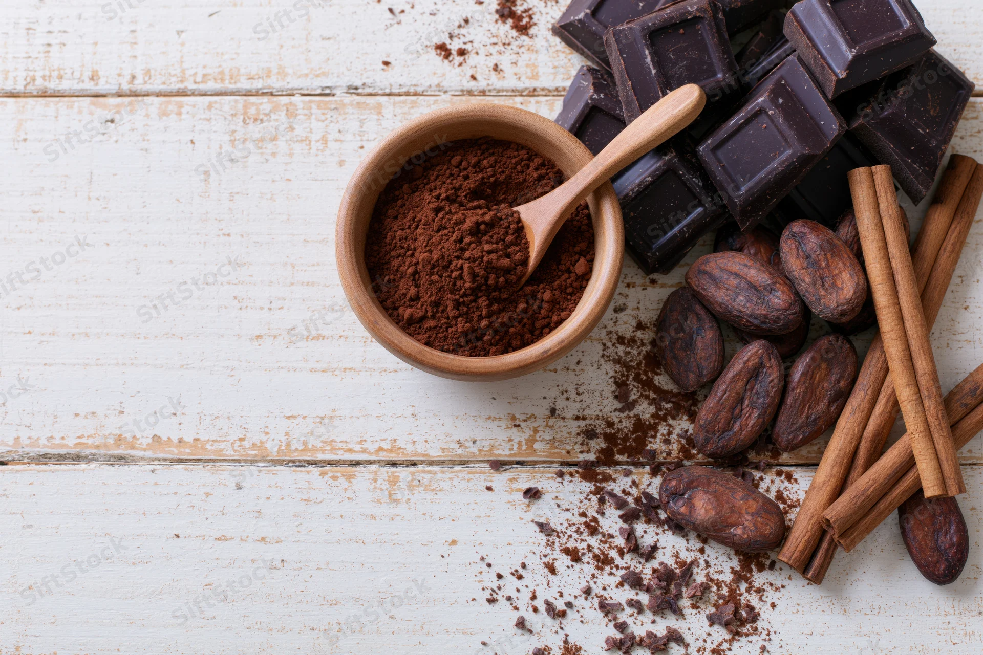 Dark chocolate chunks and cocoa powder in a wooden bowl with cinnamon sticks and cocoa beans on white wood surface