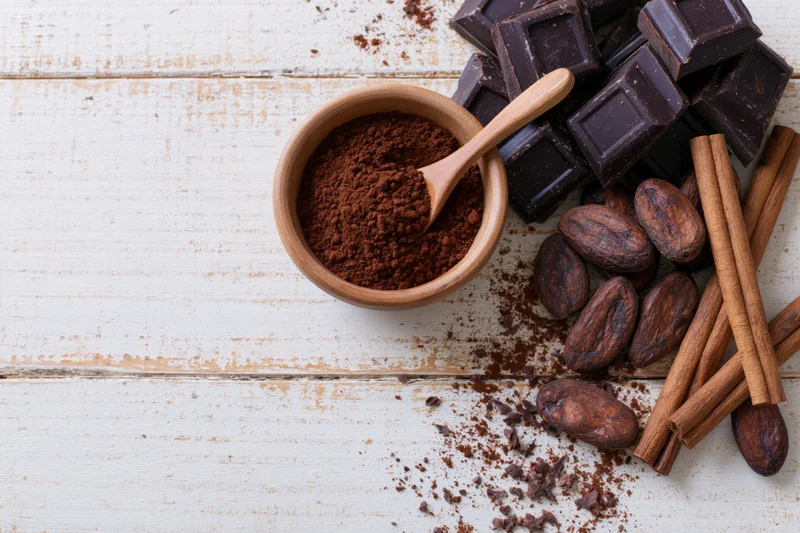 Dark chocolate chunks and cocoa powder in a wooden bowl with cinnamon sticks and cocoa beans on white wood surface