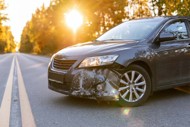 Damaged dark grey sedan with crumpled front fender on asphalt road at golden hour