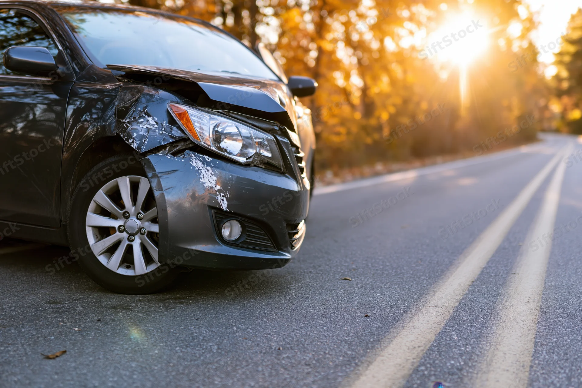 Damaged gray sedan with crumpled front fender on asphalt road at sunset