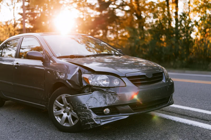 Damaged dark grey sedan with crumpled front driver-side fender on road at golden hour