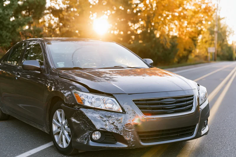Dark grey sedan with crumpled front driver-side fender parked on roadside at golden hour