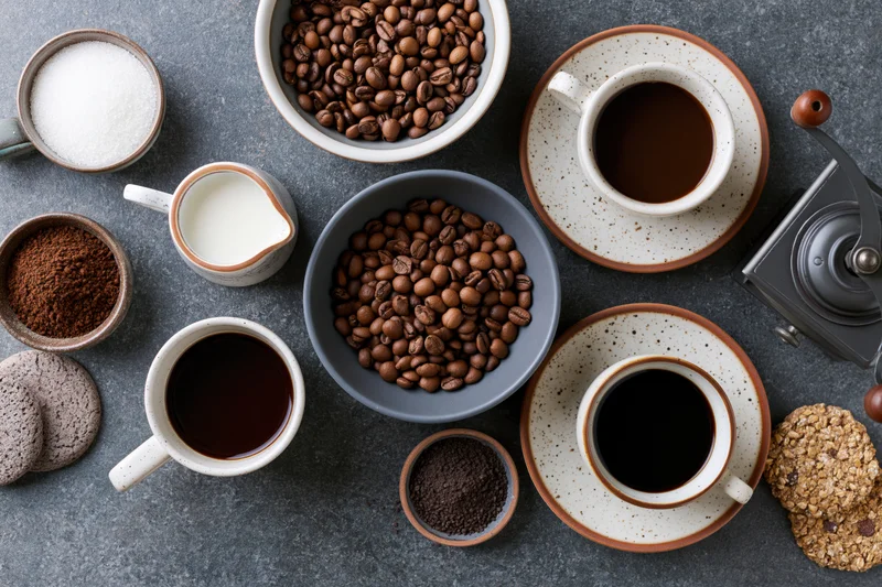 Overhead flat lay of coffee cups, beans, milk, sugar and cookies on gray surface